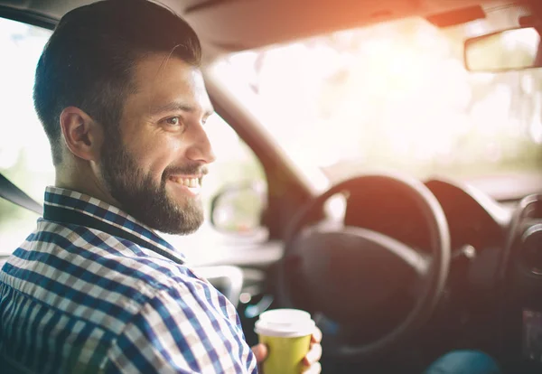 Bel homme barbu souriant assis sur les sièges passagers avant dans la voiture et elle boit du café dans une tasse jetable — Photo