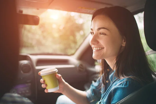 Красивая женщина улыбается, сидя на передних пассажирских сидениях в машине, и она пьет кофе из одноразовой чашки — стоковое фото