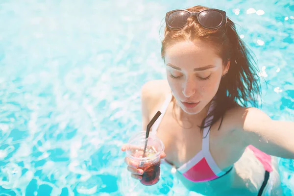 Chica morena con cócteles relajante en la piscina. Mujer sexy en bikini disfrutando del sol de verano y bronceado durante las vacaciones en la piscina con bebida. Modelo femenino hace selfi en agua — Foto de Stock