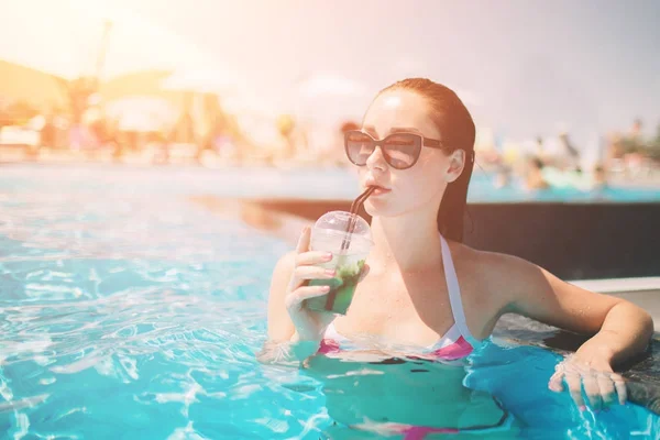 Brunette girl with cocktails relaxing in swimming pool. Sexy woman in bikini enjoying summer sun and tanning during holidays in pool with drink. — Stock Photo, Image