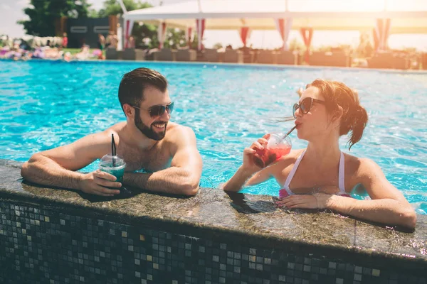 Pareja joven junto a la piscina. Hombre y mujeres bebiendo cócteles en el agua . — Foto de Stock