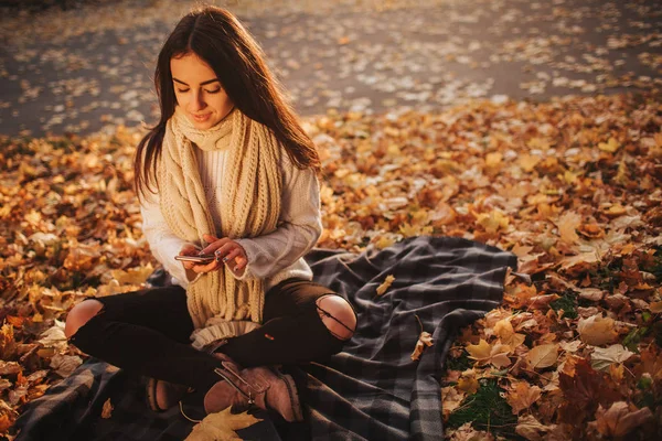 Женщина, использующая смартфон осенью. Осенняя девушка разговаривает по телефону в солнечной листве. Портрет кавказской модели в лесу осенью — стоковое фото