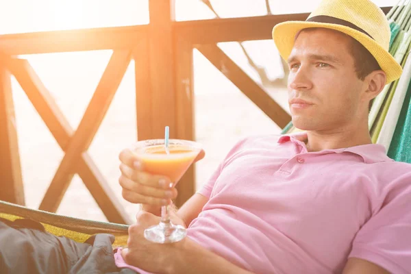 Joven guapo sosteniendo un cóctel naranja y acostado en una hamaca en una playa soleada junto a un océano — Foto de Stock