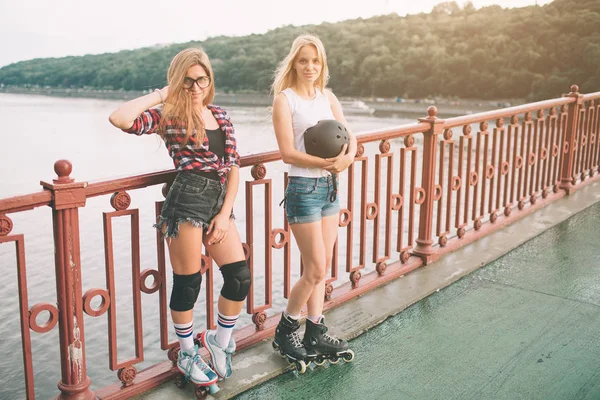 Duas mulheres jovens magras e sensuais e patins. Uma fêmea tem patins em linha e a outra tem patins quádruplos. Meninas passeio nos raios do sol — Fotografia de Stock