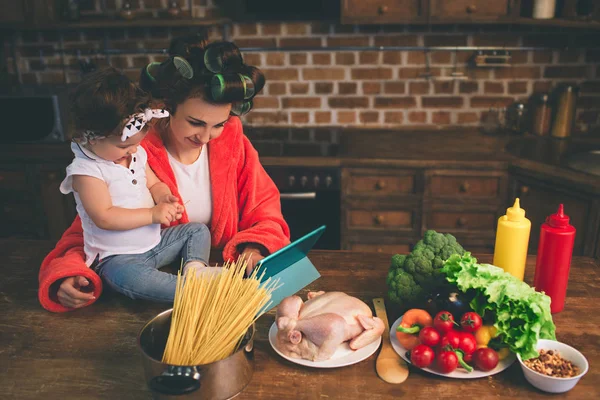 Mama acasă. Mamă tânără cu un copil mic în bucătăria casei. Femeia face multe sarcini în timp ce are grijă de copilul ei. Utilizați un PC tabletă. Tehnologie și maternitate . — Fotografie, imagine de stoc