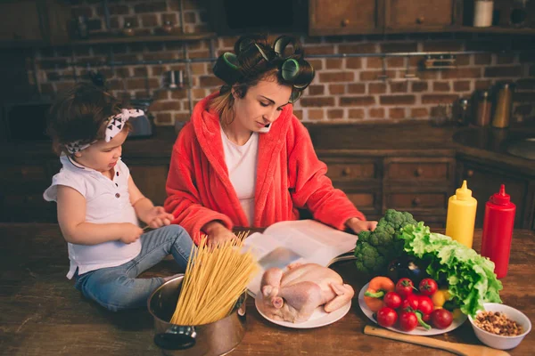 Μαμά στο σπίτι. Νεαρή μητέρα με μικρό παιδί στην κουζίνα στο σπίτι. Γυναίκα κάνει πολλές εργασίες ενώ φαίνεται μετά από το μωρό της. Διαβάζοντας ένα περιοδικό γυαλιστερό και ψάχνει για νόστιμες συνταγές — Φωτογραφία Αρχείου