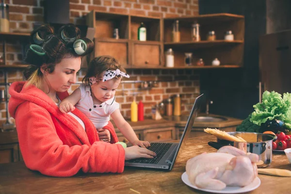 Anya otthon van. Fiatal anya kisgyerekkel az otthoni konyhában. Nő csinál sok feladatot, miközben vigyáz a baba. Laptopot használ PC technológia és az anyaság — Stock Fotó
