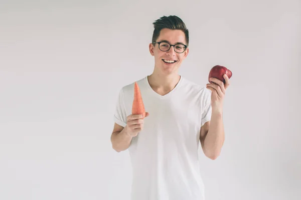 Ein Mann in weißem Hemd isoliert auf hellem Hintergrund mit Karotte und Apfel. Vegetarier bereiten eine Mahlzeit zu. lächelnder Kerl liebt Obst und Gemüse.. Nerd trägt Brille. — Stockfoto