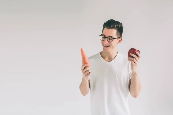 Ein Mann in weißem Hemd isoliert auf hellem Hintergrund mit Karotte und Apfel. Vegetarier bereiten eine Mahlzeit zu. lächelnder Kerl liebt Obst und Gemüse.. Nerd trägt Brille. — Stockfoto