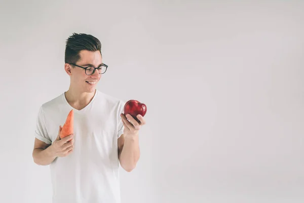 Ein Mann in weißem Hemd isoliert auf hellem Hintergrund mit Karotte und Apfel. Vegetarier bereiten eine Mahlzeit zu. lächelnder Kerl liebt Obst und Gemüse.. Nerd trägt Brille. — Stockfoto