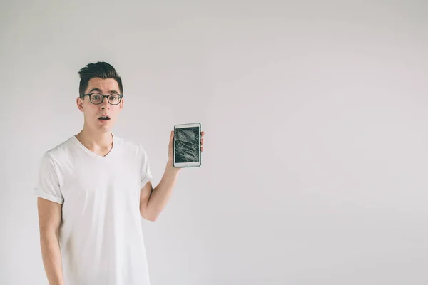 Nerd is wearing glasses. Student presenting a broken black tablet behind glass. upset man holds a out-of-use tablet or smartphone. Isolated on a light background. Broken screen