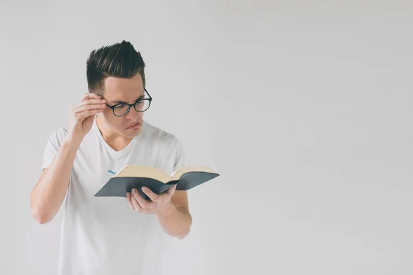 Nerd con occhiali e una t-shirt bianca sta leggendo un libro su uno sfondo bianco — Foto Stock