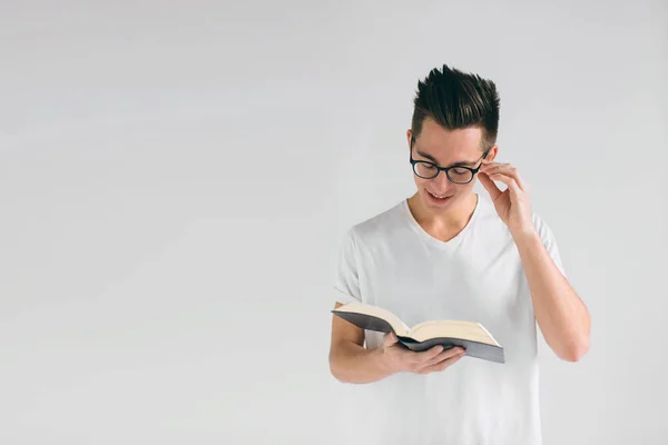 Nerd con occhiali e una t-shirt bianca sta leggendo un libro su uno sfondo bianco — Foto Stock