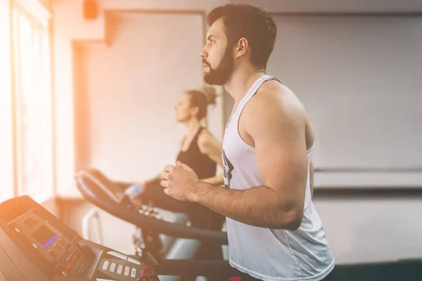 Jovem de fato desportivo a correr em esteira no ginásio. Atleta barbudo muscular durante o treino. Close up de jovens atléticas modelo feminino trens no centro de fitness indoor . — Fotografia de Stock