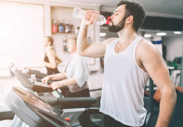 Giovanotto in abbigliamento sportivo che corre sul tapis roulant e beve acqua in palestra. Atleta muscoloso barbuto durante l'allenamento. Primo piano di giovani allenamenti sportivi femminili presso il centro fitness al coperto . — Foto Stock