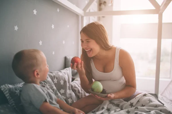 Várandós anya és a fia kis fiú eszik egy alma és az őszibarack a bed-t otthon reggel. Alkalmi életmód a hálószobában. — Stock Fotó