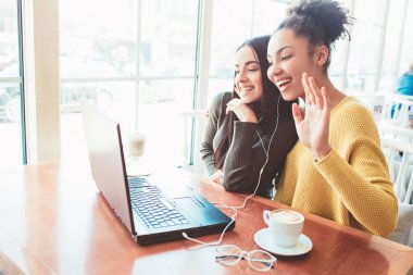 Girls are sitting in cafe and chatting with somebody . Probably its their third friend that cant be with them. Young women are waving to their friend that is on the other side of the screen clipart