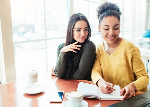 Studenten zitten in de buurt van het venster en studeren. Het meisje in het geel maakt sommige nota's in haar notitieblok, terwijl het meisje in bruin is op zoek naar laptops scherm. Jonge vrouwen zijn voorbereiding voor examen. — Stockfoto