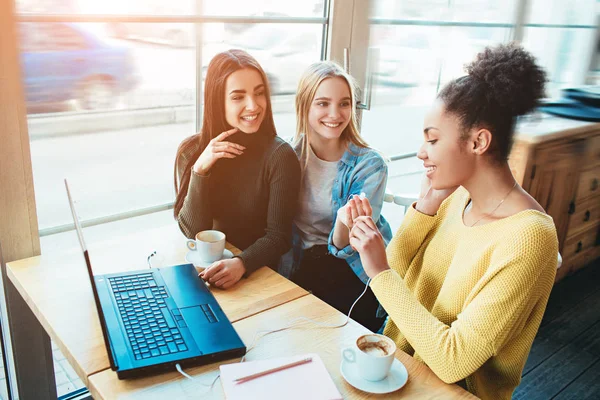 Árbol de las mujeres jóvenes están sentados juntos en un pequeño café con grandes ventanas y hablando entre sí. Uno de ellos está escuchando música y es muy feliz y dos simplemente disfrutando de su conversación — Foto de Stock