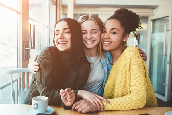 Boom jonge vrouwen zitten samen in een klein café met grote ramen en omhelzing met elkaar. — Stockfoto