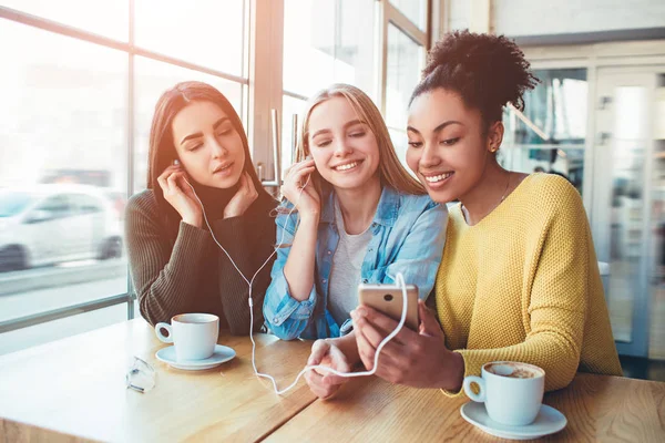 Drei junge Frauen, die lächeln. Sie schlossen die Augen und genossen die Musik auf dem Smartphone. beste Freunde sitzen im Café — Stockfoto
