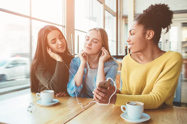 Drie jonge vrouwen die zijn glimlachen. Ze sloot haar ogen en genieten van de muziek op de smartphone. Beste vrienden zitten in café — Stockfoto