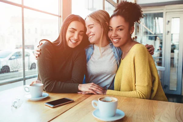 Boom jonge vrouwen zitten samen in een klein café met grote ramen en omhelzing met elkaar. — Stockfoto