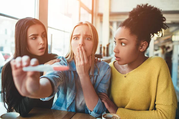 blonde girl is holding a pregnancy test that shows to her positive result of the test. She looks dissapointed, sad and trying not to cry. Her two best friends are trying to support her