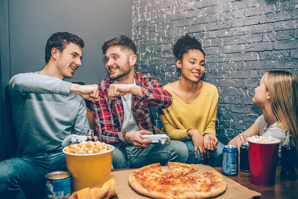 Vänner sitter runt bordet i grå rum och spendera lite tid tillsammans. Pojkar hälsning varandra medan flickor pratar och ha lite kul. De förberedda välsmakande pizza. Gamers. Night part. — Stockfoto