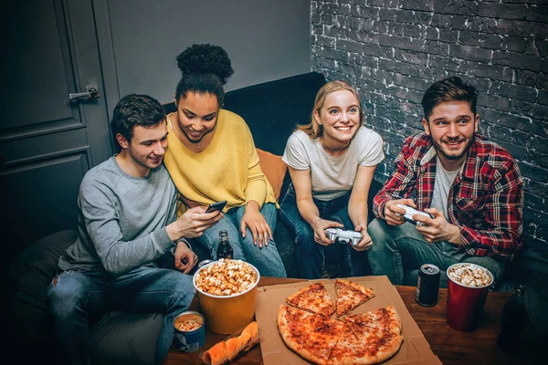Two teenagers are looking to the smartfone and laughing while the other two are playing game using gaming console. All of the have a lot of fun. Gamers. Night party.