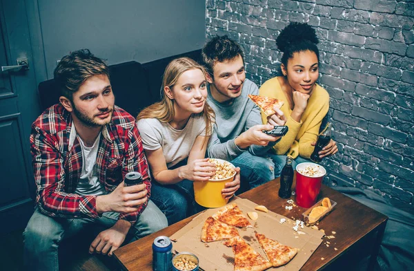 Excited young people are sitting together on a couch, eating popcorn and watching different movies. They are concentrated only on that. — Stock Photo, Image