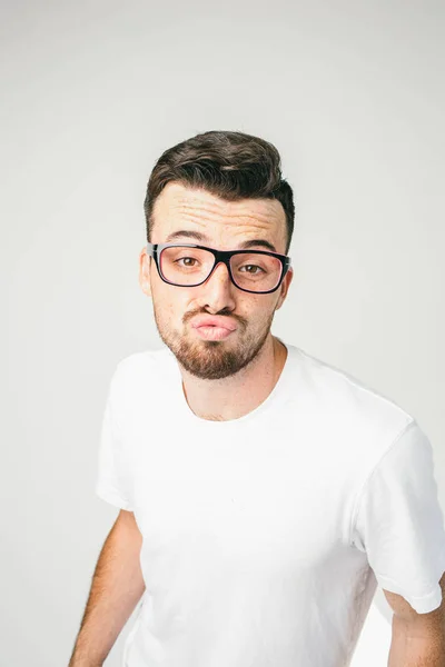 A vertical portrait of a dark-haired and bearded man standing at white wall wears glasses and looking straight to the camera. He has very strange and arrogant sight. Close up. Cut view — Stock Photo, Image