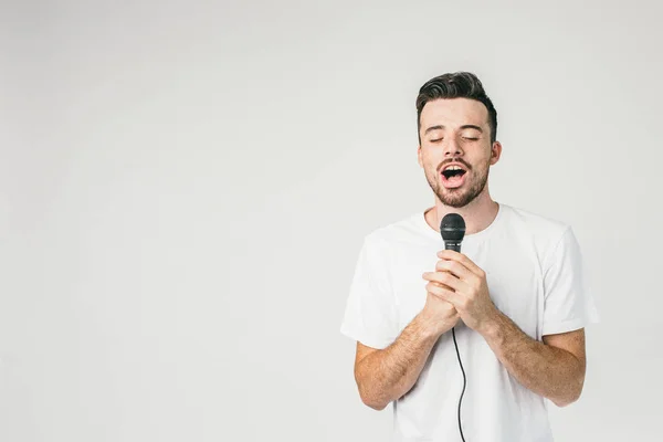 Un tipo valiente parado en la pared blanca y sosteniendo un micrófono con sus dos manos. Está cantando una canción sensual y triste con los ojos cerrados . —  Fotos de Stock