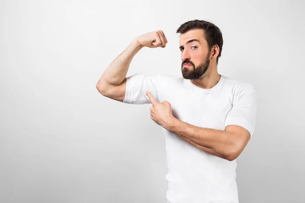 A picture of dark-haired and bearded guy standing near white wall and pointing at his big muscles on his right hand and looking to camera. This guy is confident he has good body shape. — Stock Photo, Image