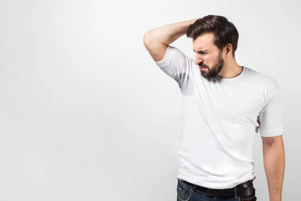 A picture of handsome guy is sniffing his armpit. There is a big sweat spot and the guy doesnt like it at all and shrinking. He doesnt like the smell of it. Isolated on white background. — Stock Photo, Image