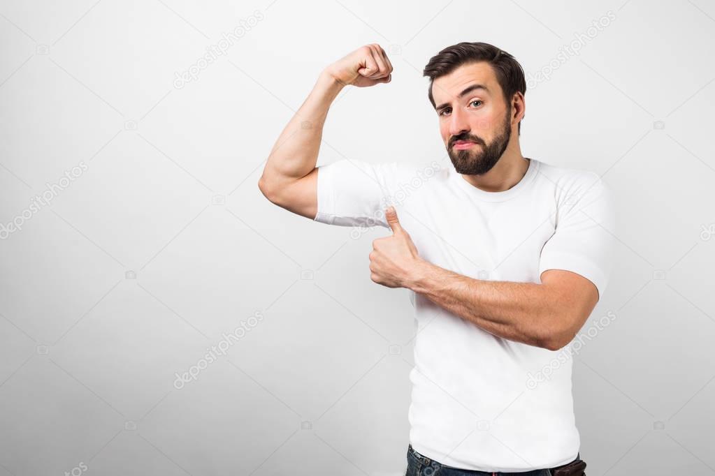 Young man standing isolated on white background in white shirt and showing a big thumb up in front of his arm muscles. Its because his body is in a good shape and the guy knows about that. background