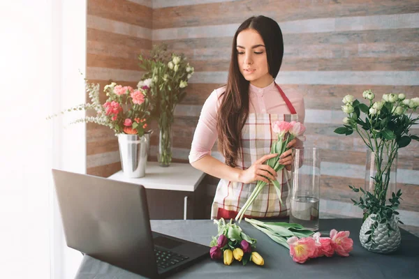 Female florist at work: pretty young dark-haired woman making fashion modern bouquet of different flowers. Women working with flowers in workshop. She is using the laptop at work