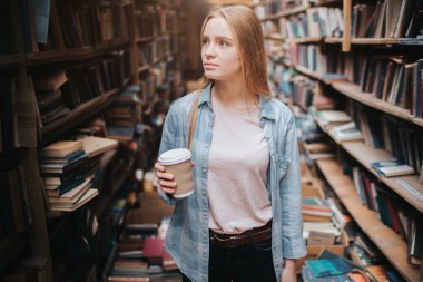 Çekici ve güzel bir kız eski kitaplar ile büyük ve uzun bookshelfs arasında duruyor. O kahve bir fincan elinde tutan ve raf için arıyorum. Kadın bir kitap için arıyor.