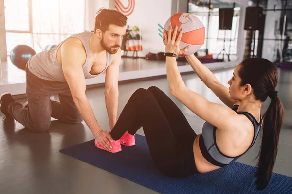 Una foto di una ragazza che fa un po 'di esercizio addominali con la palla mentre il suo partner sportivo sta tenendo le gambe sul pavimento. Lui la aiuta a fare esercizio in modo corretto . — Foto Stock