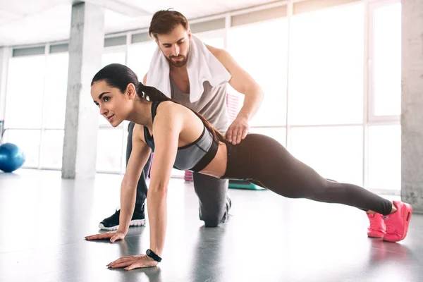 El entrenador y su estudiante están en una sala de fitness. Ella está haciendo ejercicio de tabla alta mientras él controla la calidad de lo que ella está haciendo . — Foto de Stock