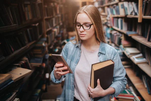 A girl standing near long wall full of bookshelf with different books. She is holding a book in her left hand and a smartphone ith the right one. She is looking to the screen. — Stock Photo, Image