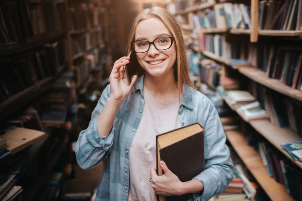 A picture of attractive girl standing with a book in her left hand and talking on the phone. She is looking straight forward and smiling. This woman is very nice. — Stock Photo, Image
