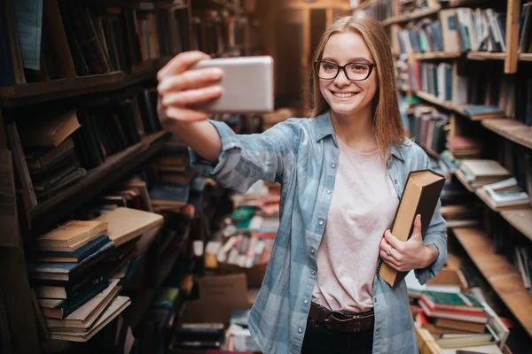 A cute picture of a blonde girl taking selfie. She is looking to the phone and smiling. This girls is in a big public library. — Stock Photo, Image