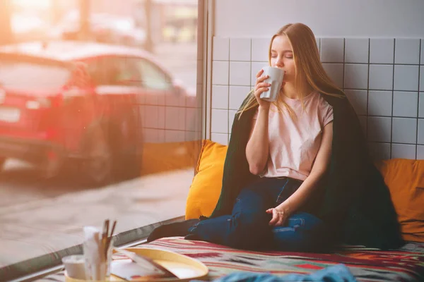 Krásná roztomilá dívka v kavárně sedí na kolenou u okna s šálkem kávy a při pohledu na ulici. Vypadá thoughtfl a klid. — Stock fotografie