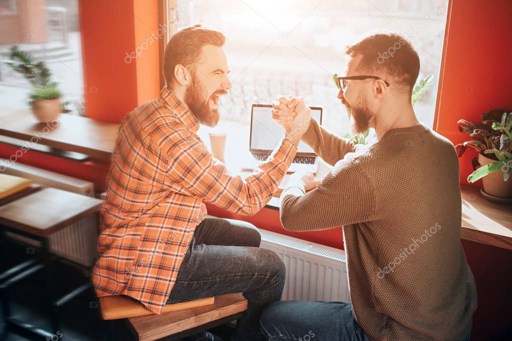 Nice and good picture of two guys sitting at the table in front of the big window and holding each other with that fists. THey are congratulating each other