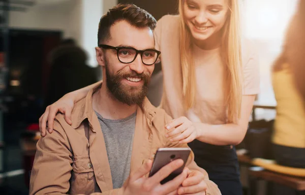 Close up and cut view of a bearded man in jacket sitting in cozy cafe at the table and holding a phone in his hand. The young girl stands beside him and looking down to the phone.