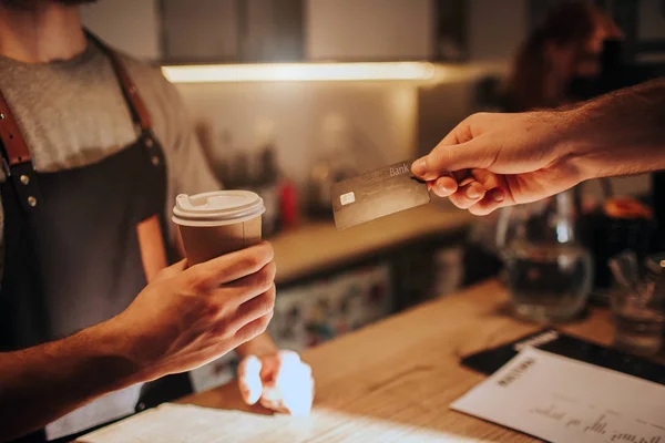 Kadar yakın ve bir fincan kahve tutarak ve müşteriye veren bir barmen el görünümünü kes. Aynı zamanda müşteri ona sipariş için ödemek için bir kredi kartı veriyor. — Stok fotoğraf