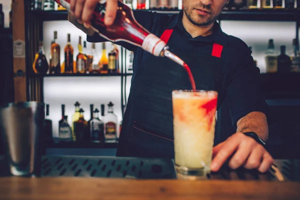 Barman adicionando xarope vermelho no copo cheio de bebida alcoólica branca. O líquido está a misturar-se no vidro. Homem está segurando o vidro com dois dedos . — Fotografia de Stock