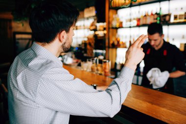 Barmen doğru oturan müşteri resmi. Kokteyl ve sağ eliyle gösterilen sipariş etmek istiyor. Barmen bunu görmüyor.
