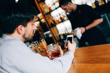 Barmans standında oturmuş güzel ve kaslı adam. O sağ eli ve sol bir içki cam fincan bir telefon tutun. Konsantre ve ciddi görünüyor. Barmen yakın duruyor.